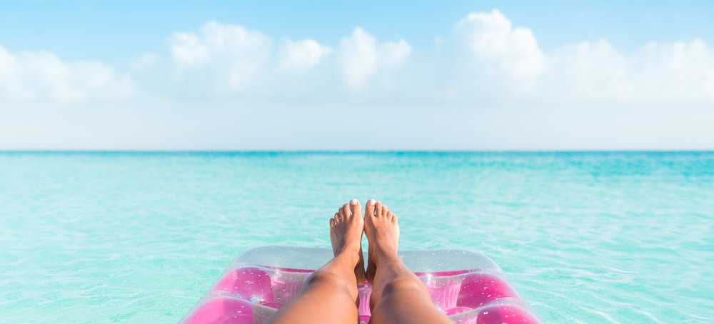 womans feet on a pink raft floating in pale blue water